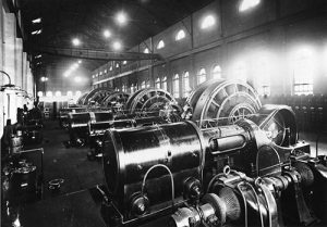 An interior view of the Denver Tramway Company power plant (now REI) at 14th and Platte. A line of large turbines fill the room and one wall is lined with electric switches. Image courtesy Stephen H. Hart Library & Research Center, History Colorado.