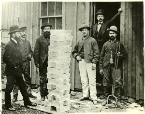 a group of men standing around a stack of silver bullion bars (ingots) at the Argo Smelting Company in Black Hawk, Colo., photographed by an unidentified photographer circa 1873. A caption in the album indicates that this was the first silver made by Richard Pearce (b. 1838; Manager at Argo) and that Pearce "brought these men to Colorado with him from Swansea, Wales." Men are identified as Richard Pearce (on left), Josiah Burgess (refiner; on right), Thomas Thomas (mason; in light overalls), and William Abbe (foreman; in doorway).